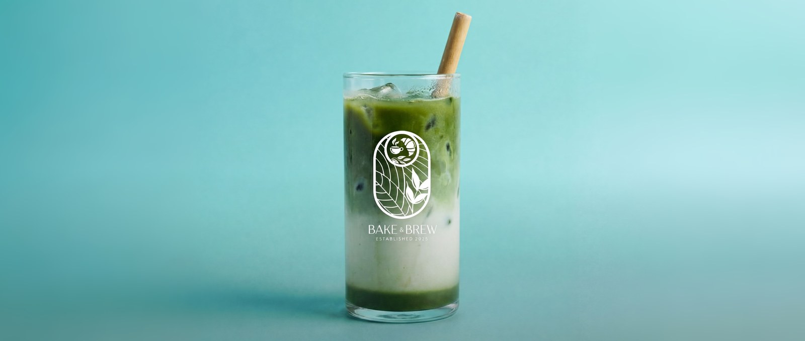 Eiskalter Matcha Latte von Bake & Brew in einem hohen Glas mit dem eingravierten Bake & Brew Logo und einem Bambus-Strohhalm, vor türkisblauem Hintergrund.